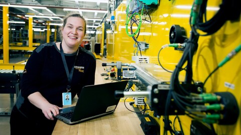 Amazon Appretice, Nicola Elliott, leaning at her workbench with with her laptop. She is in the Bolton Fulfilment Centre next to a yellow wall with lots of wires and equipment attached to it.