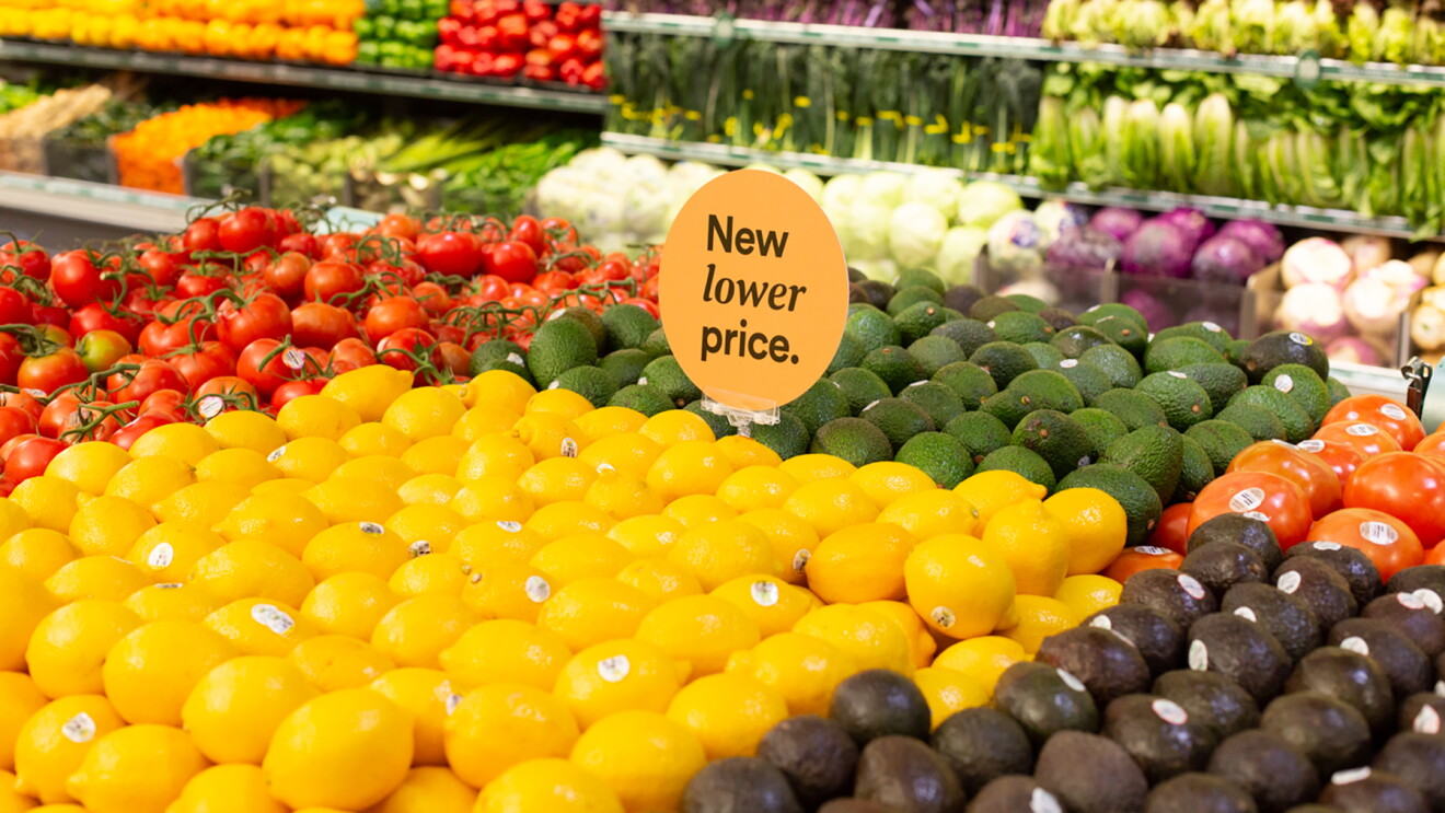 Inside a Whole Foods Market store, lemons, limes, avocados, and tomatoes are stacked on a table, with a "New lower price" sign in the center of the produce. Behind the display, lettuces, peppers, squash and other vegetables are stocked in a produce wall display. 