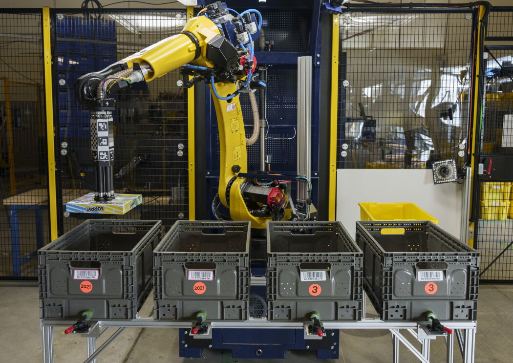 Il nuovo braccio robotico Sparrow, immortalato in diverse posizioni, solleva dei piccoli oggetti da quattro contenitori di plastica grigia allineati davanti a lui