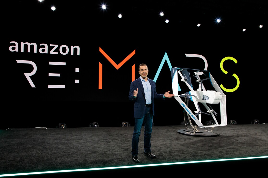 Jeff Wilke, CEO consumer worldwide at Amazon, stands on a stage. Behind him is a re:MARS logo. To his left is a drone painted in white and light blue. 