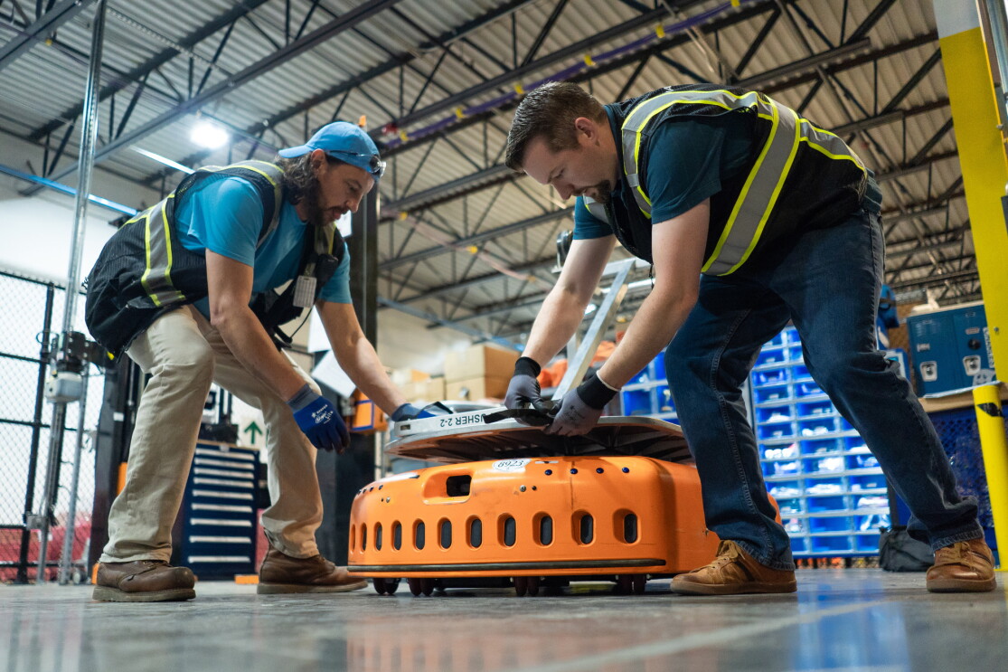 Dentro de un centro logístico con las columnas de estanterías detrás, aparecen dos hombres arreglando un robot de Amazon. Los dos hombres van con un chaleco de color negro con rayas amarillas. El robot de color naranja está en el centro. De los dos hombres, el de la izquierda va con una gorra de color azul y guantes de seguridad. Su compañero de la izquierda también va con guantes. 
