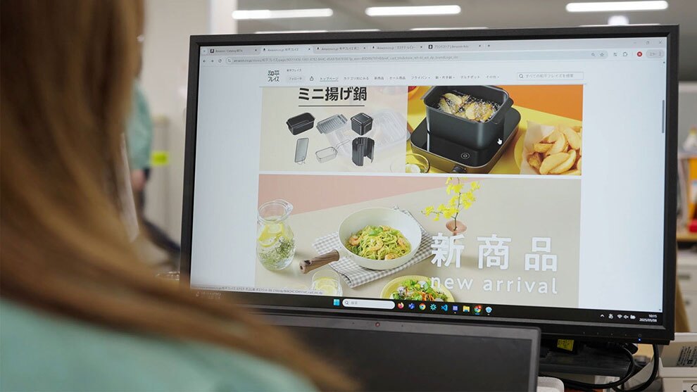 モニターに映された商品販売サイトの画面。ミニ揚げ鍋の商品画像とその下に「新商品」の文字が表示されている。左手前にモニターを見る人物の後ろ姿があるが、ピントは画面に合っている。