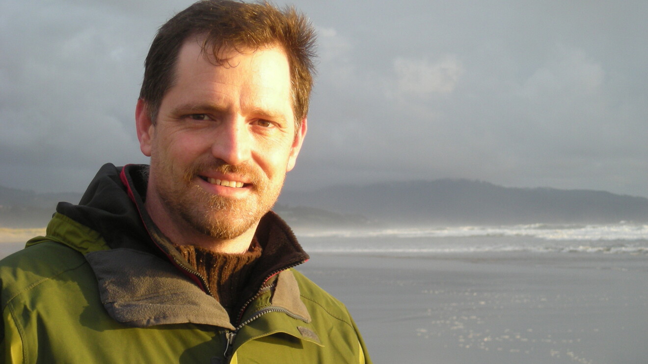 A man in a green jacket standing on the beach in high surf.