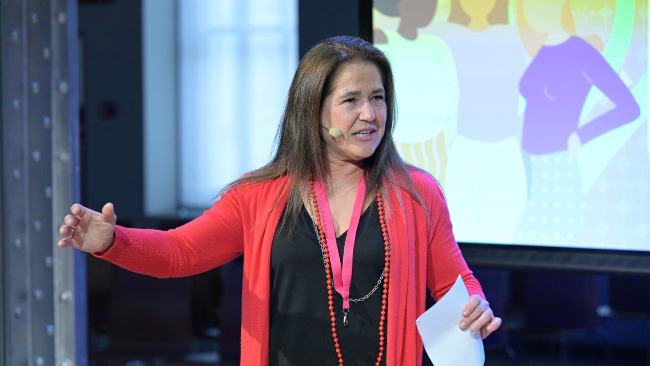 María Gómez del Pozuelo, cofundadora y CEO de Womenalia. Aparece de pie con un vestido negro, una chaqueta de color rojo y un collar largo por la cintura de perlas rojo. De fondo se ve una pantalla con la silueta dibujada de tres mujeres que se abrazan. María está hablando con un micrófono en la mejilla, un papel en la mano izquierda y con la mano derecha está gesticulando.