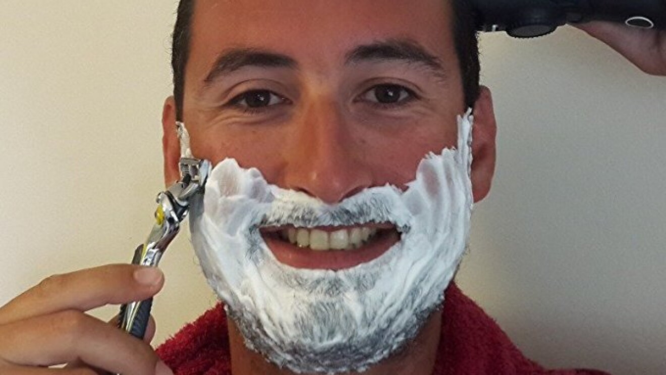 Un homme torse nu avec une serviette autour du cou a la barbe couverte de mousse à raser et tient dans chacune de ses mains un rasoir mécanique et un rasoir électrique. Il les tient posés sur ses cheveux et sa barbe en souriant.