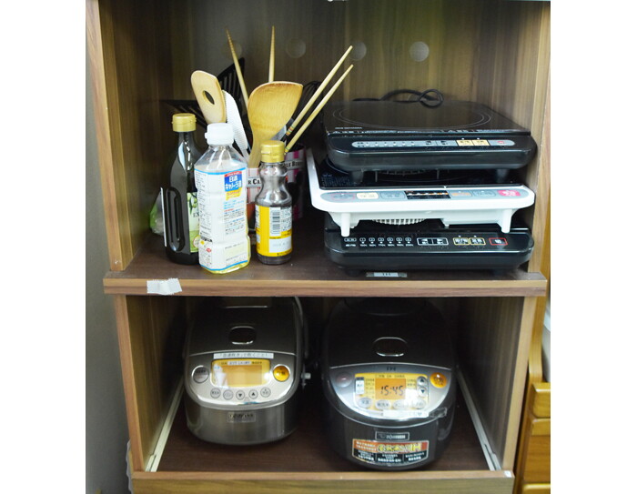 茶色い棚の二段。上段にホットプレート3台と調理器具や油類、下段に炊飯器2台。
