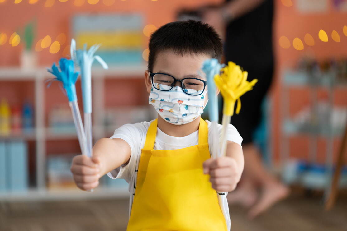 A young boy holding up his artwork, wearing a yellow apron