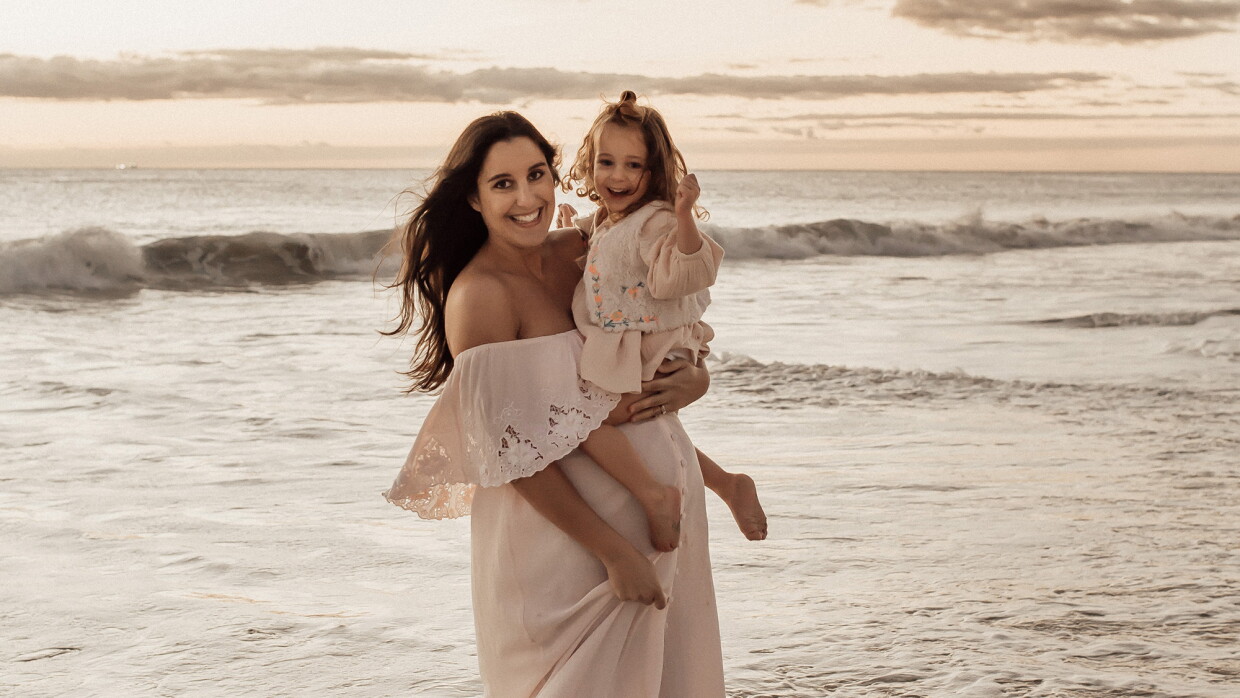 A woman stands on a beach wearing a white flowing dress, smiling for a photo while holding her child. 