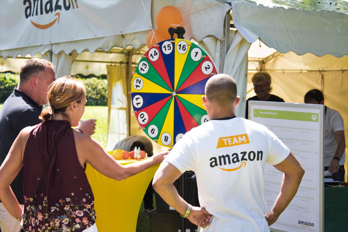 Ein Amazon Mitarbeiter steht vor einem bunten Glücksrad. Er ist von hinten zu sehen. Auf dem Shirt ist "Team Amazon" zu lesen.