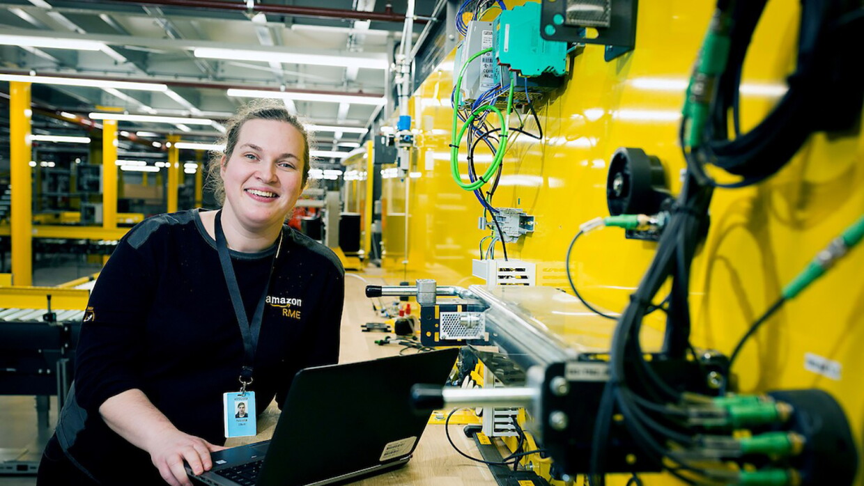 Amazon Appretice, Nicola Elliott, leaning at her workbench with with her laptop. She is in the Bolton Fulfilment Centre next to a yellow wall with lots of wires and equipment attached to it. 