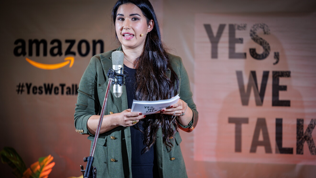 En un escenario creado para la ocasión, de fondo hay un cartel con el logo de Amazon, el Yes, we talk, que es el lema de la campaña y el lema de la sesión Rompiendo el silencio. Todo el fondo es de color rosa. En el escenario, 6 plantas con sus respectivas macetas, dos mesas y dos sillas. Nicole, una trabajadora de Amazon, está de pie delante de un micro y hablando agarrando con las dos manos unos papeles. Sentada en la silla y mirando a Laura está la directora de cine Leticia Dolera. Nicole va vestida con un vestido negro por la rodilla y una americana verde militar. Unos zapatos de tacones animal print. Con un pantalón acampanado, un jersey y una americana. Lleva el pelo largo, ondulado, negro hasta la cintura. Leticia Dolera va con una americana gris, jersey negro de lana de cuello alto, pantalón negro y botines animal print. Es pelirroja, lleva el pelo largo hasta el pecho y todo a un lado. Leticia está mirando a Nicole.