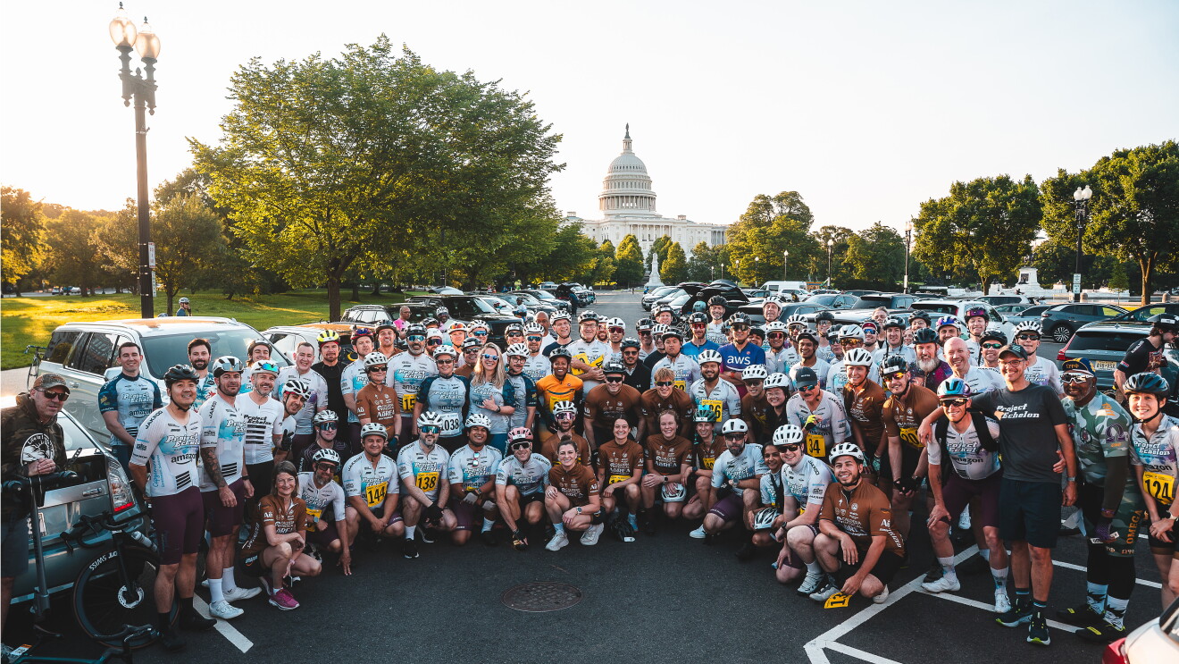Large group of cyclists in matching team jerseys posing together in a parking lot with the U.S. Capitol building visible in the background