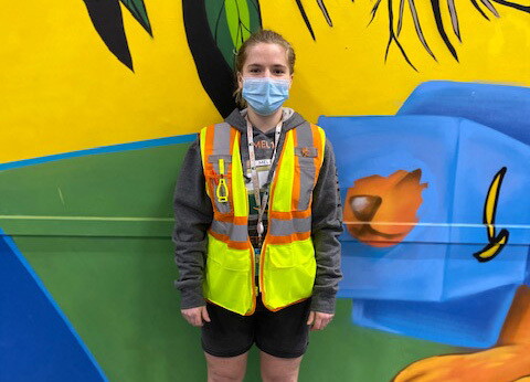 An image of Lisa smiling for a photo (with a mask on) while standing in front of a bright-colored wall in the fulfillment center where she works. She is wearing a neon safety vest. 