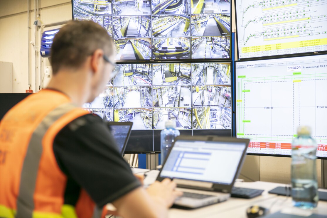 Ein Mitarbeiter in orange-farbenen Weste tippt an einem Laptop. Vor ihm sieht man auf großen Bildschirmen Technik und Förderbänder.