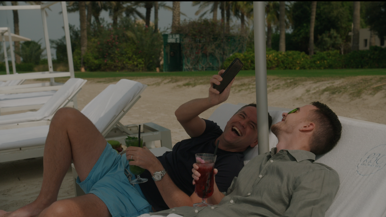 Michael Owen and son lie on loungers on the beach with a cocktail in hand, laughing at something on Michael's phone screen