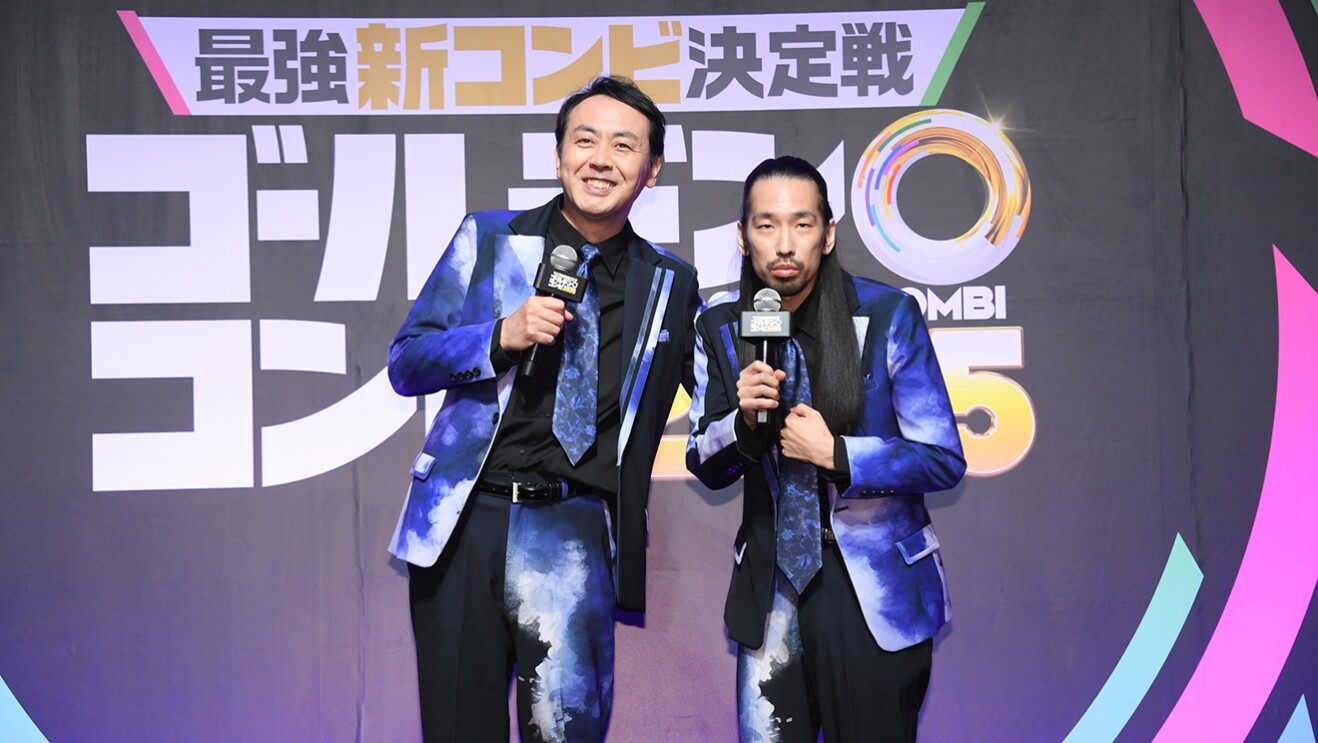 黒地に白の大きな文字が書かれた壁を背に、マイクを持って立つ2人の男性。2人とも黒と紺色マーブルの派手なスーツ衣装を着用。
