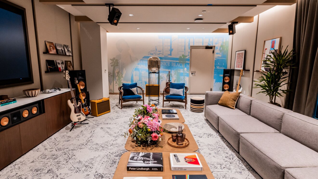 A green room with a couch, two blue chairs, and a large table with flowers and coffee table books on it. There are several big speakers located throughout the room, and a guitar displayed in front of the TV stand. 