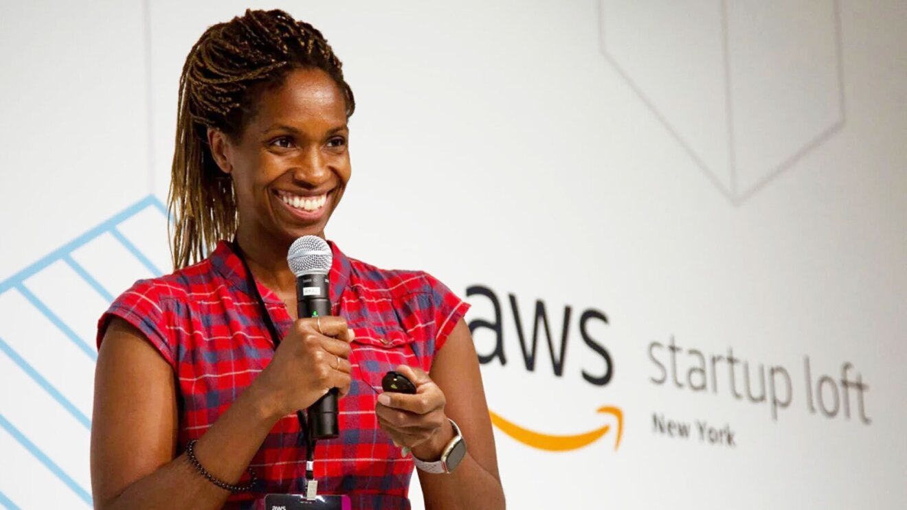 Alyse stands on a stage smiling while holding a microphone. There is a background behind her that reads "AWS startup loft, New York"