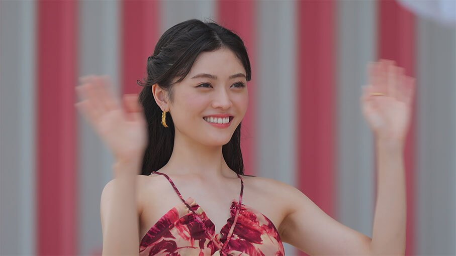 紅白の縦じまの布を背景に、笑顔の女性が肩の上あたりまで上げた両手を振っている。