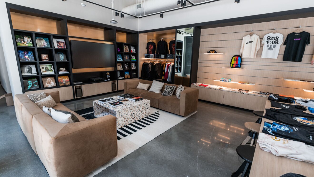 A living room area with two couches and a table in between them. There is a wall with record covers on it and next to it a wall with various artist merchandise like shirts and sweaters. There is also a TV on the wall.