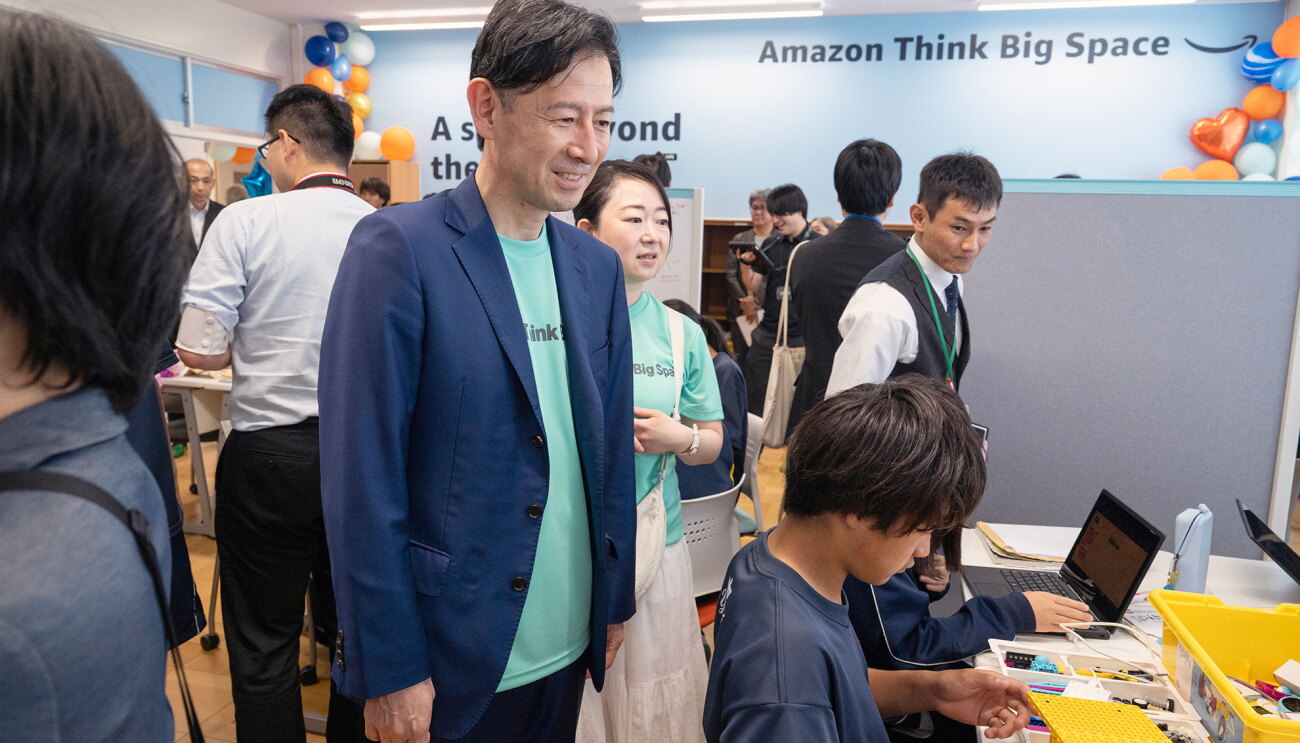 青い壁に「Amazon Think Big Space」と書かれた教室。ミントグリーンのTシャツに紺のジャケットを羽織った人物が、同じTシャツを着たスタッフと共に、作業に取り組む生徒の様子を見ている。作業机の上には、ノートPCと黄色い部品ボックス。教室を見て回る大人が他にも数5人ほどいる。