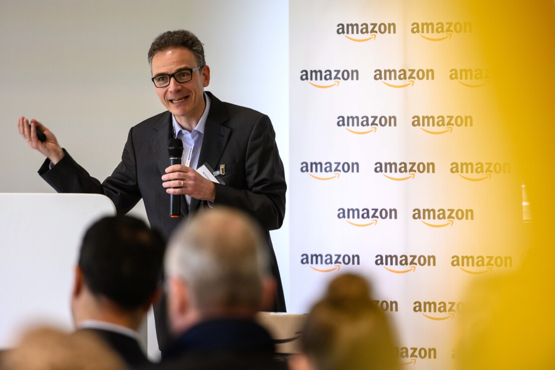 Ein Mann mit Mikrofon in der Hand, im Hintergrund sieht man eine Logo-Wand mit Aufschrift "Amazon"