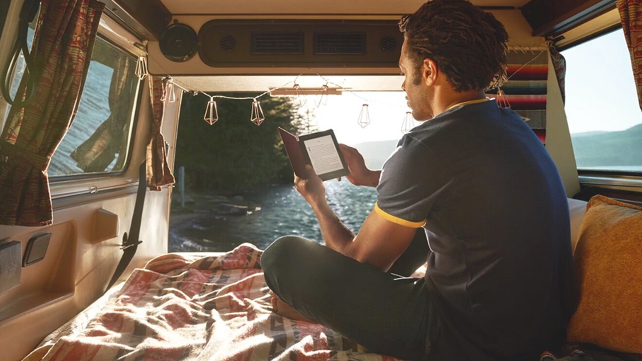 A man sits in the back of a camper van, in front of a large body of water. He is holding a Kindle Paperwhite and appears to be reading.