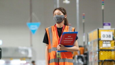 A woman wearing an orange safety vest and a face mask stands in an Amazon fulfillment center, carrying a clipboard that says "safe distancing ambassador." 