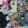 Ana Gallardo regente El Detallito, una papelería y agencia de viajes en Los Corrales, Sevilla. En la papelería Ana entrega un paquete a un cliente. Los dos llevan mascarilla. Ana está detrás del mostrados lleno de libros. En el fondo estanterías llenas de productos de papelería. Ana va con una chaqueta gris, media melena y una mascarilla negra. El cliente va con un anorak negro y una mascarilla blanca y tiene el pelo corto negro. 