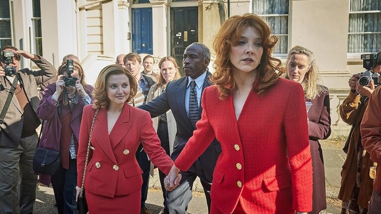 Two women wearing tailored red suits hold hands, with paparazzi in the background