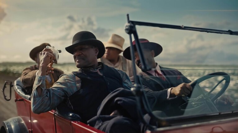 Scene featuring men in 1920s attire riding in a red convertible car