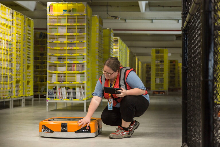 Foto di Maria Stella Carnà che regge un tablet di controllo e si china su uno dei robot di cui è responsabile all'interno del Centro di Distribuzione Amazon di Passo Corese