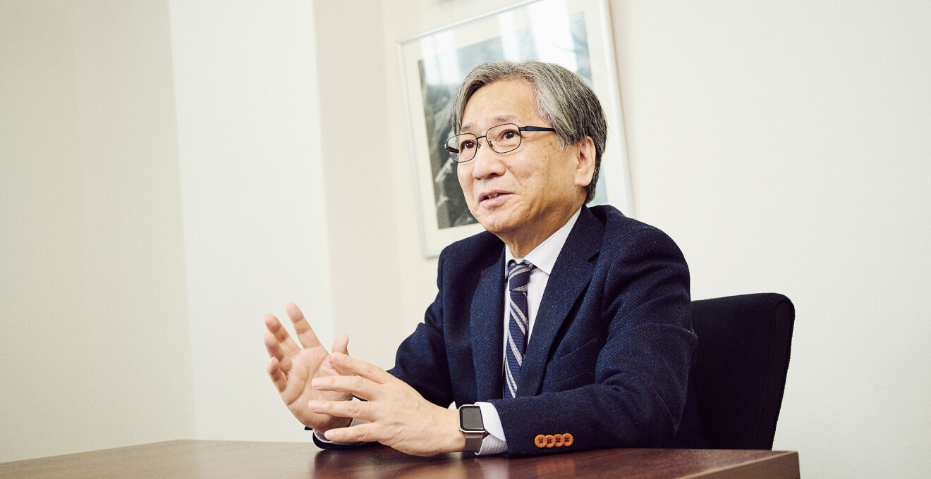 会議室のような部屋で椅子に座り話をするメガネをかけたスーツ姿の男性