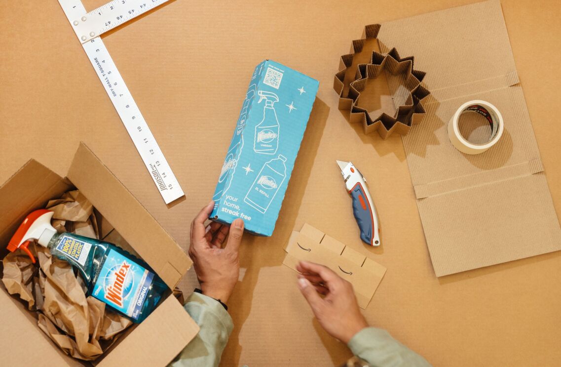 A hand holding sky-blue cardboard with spray bottle and plastic bottle illustration on it. On the left side, there is a bottle of Windex spray inside a cardboard box.