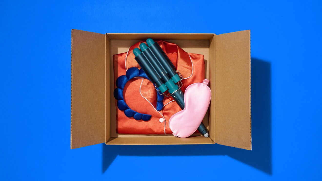 Open cardboard box containing orange folded clothing, blue water gun, braided blue rope bracelet, and pink sleep mask