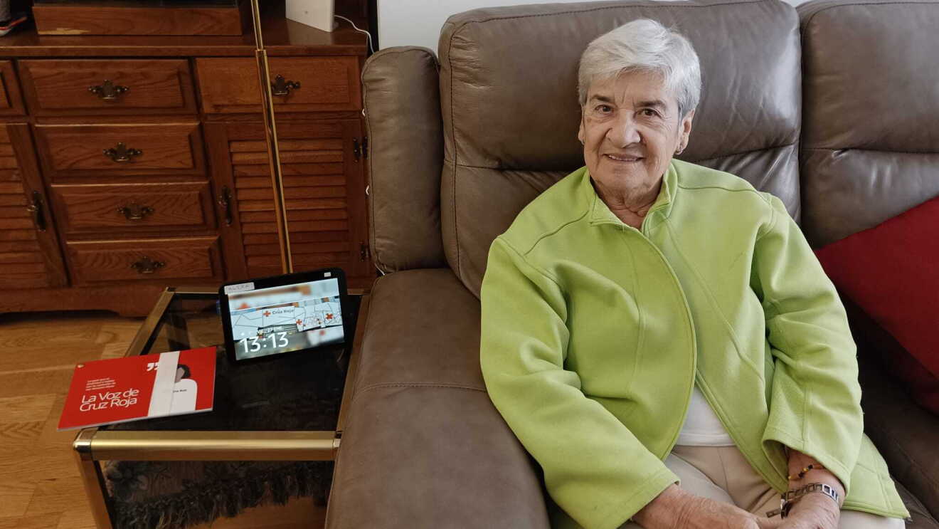Mujer mayor sentada en el sofá con una mesita con Alexa y un documento de Cruz Roja.