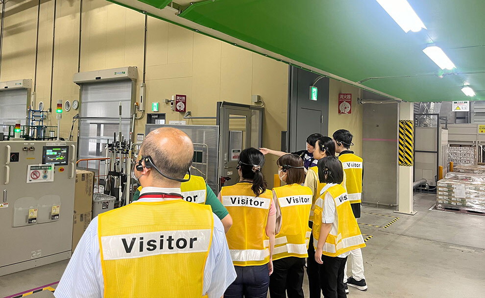 工場か倉庫の屋内でガイドの説明を聞く、7人グループの後ろ姿。7人は背中に「Visitor」の文字が付いた黄色い安全ベストを着て、黒いヘッドセットを装着している。人々の左側には傾向緑のランプやディスプレイが付いた機器、右側の緑の天井に蛍光灯。
