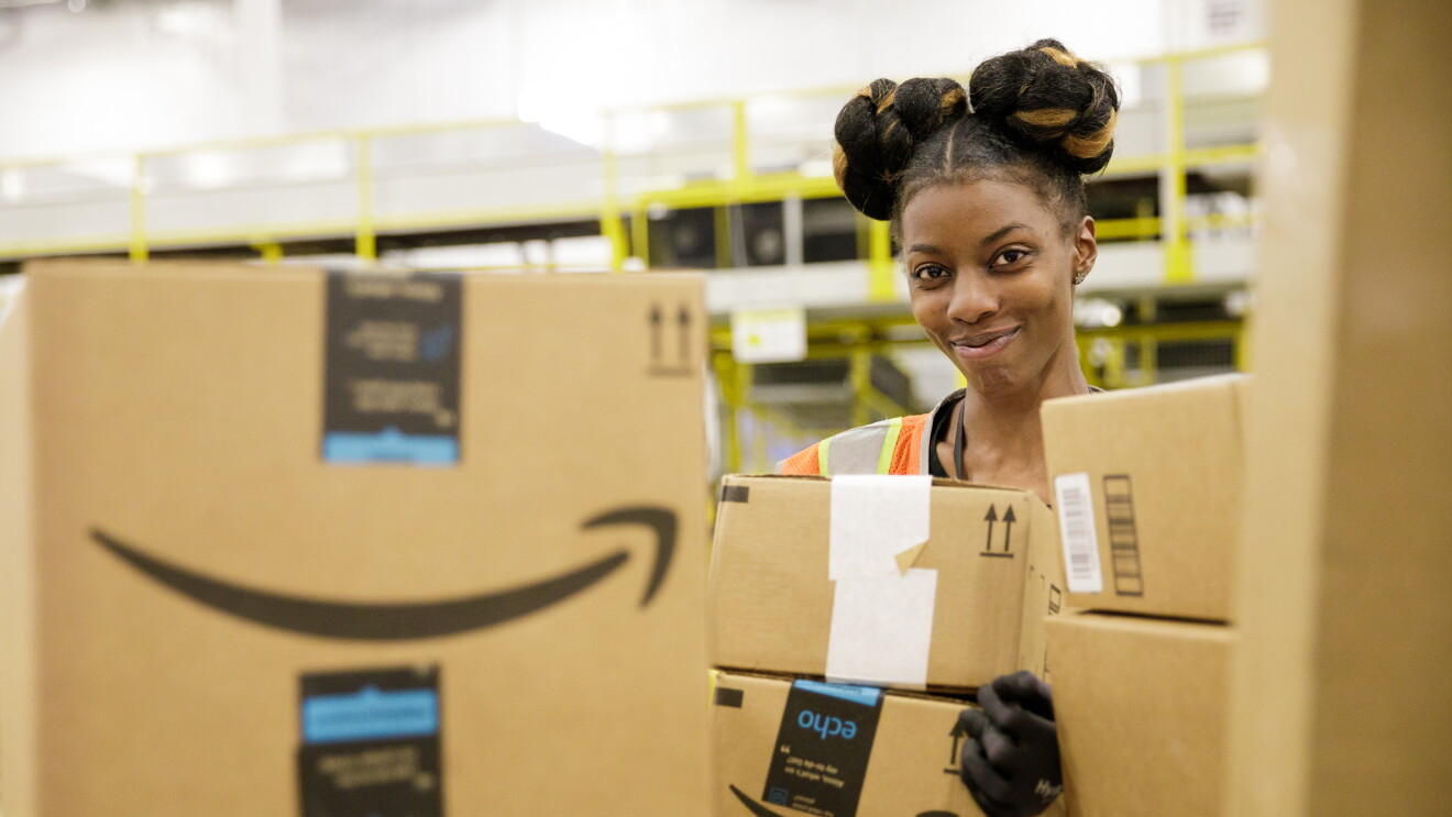 A smiling Amazon sortation associate in our Baltimore, Maryland sortation center looks at the camera from behind a pile of Amazon delivery boxes.