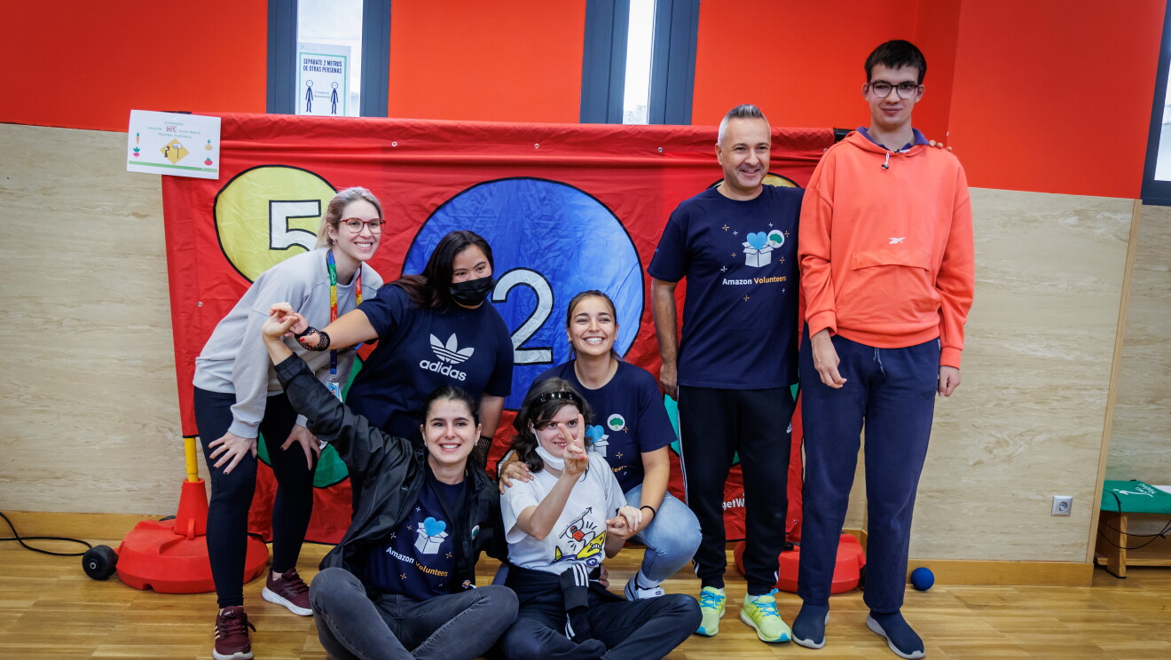 En las instalaciones de Down Madrid en Madrid aparecen 7 personas, cuatro trabajadores de Amazon y tres participantes de la fundación. Los trabajadores de Amazon van con la misma camiseta azul y el badge colgado y los de Down Madrid con deportivas y ropa cómoda. El fondo es de color rojo y el suelo de parque de pista de baloncesto cubierta.