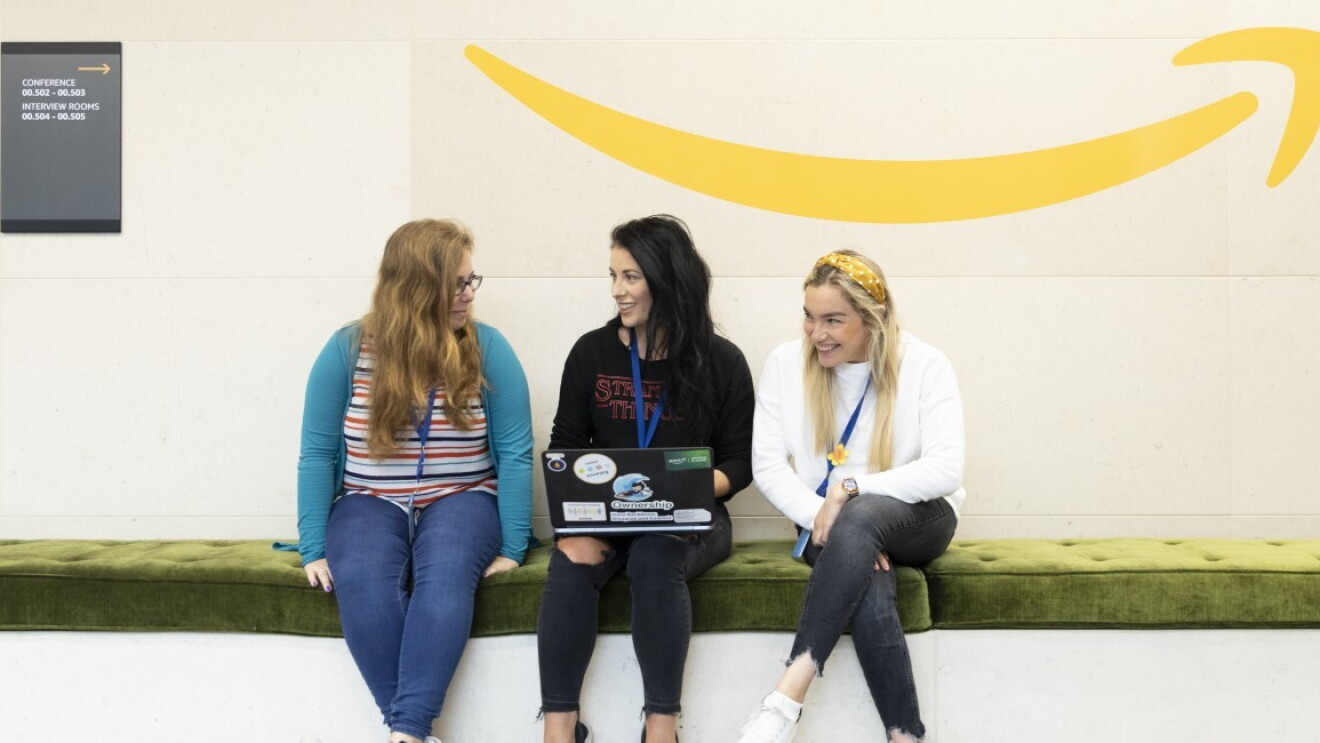 Drei junge Frauen sitzen auf einem Sofa. Sie haben einen Laptop in der Hand. Im Hintergrund sieht man das Amazon Logo.
