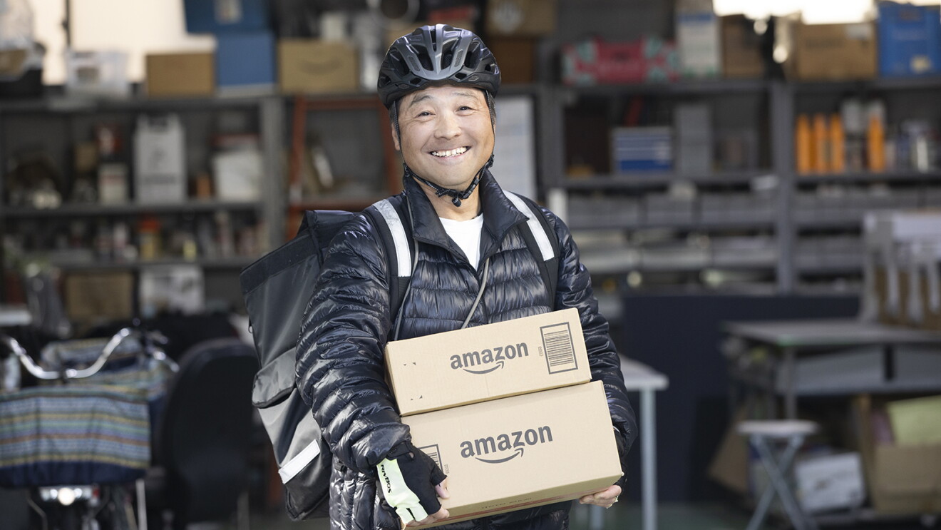 グレーの棚が並ぶガレージを背景に、自転車用ヘルメットをかぶりAmazonの段ボール箱2つを持つ男性。背中に配達用のバックパック。
