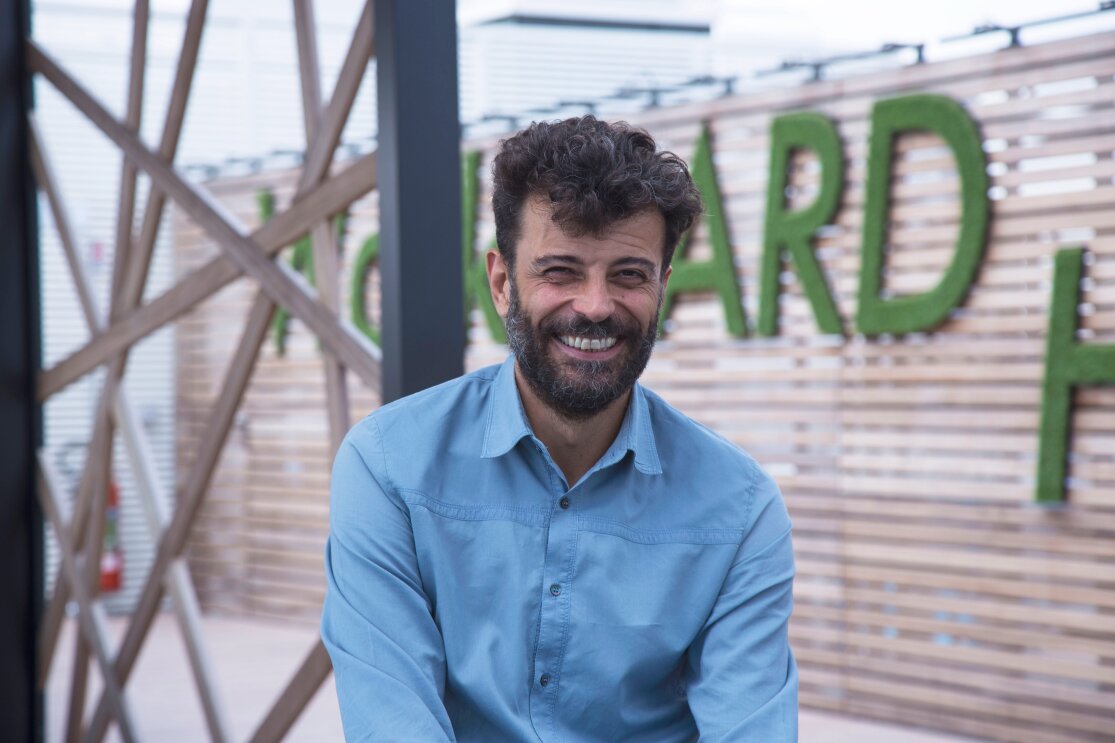 Alessandro Saccon, PR Manager di Amazon, sorride in primo sul terrazzo degli uffici Amazon di Milano. Sullo sfondo uno scorcio del terrazzo, con alcune lettere del motto 'Work hard' di Amazon. 