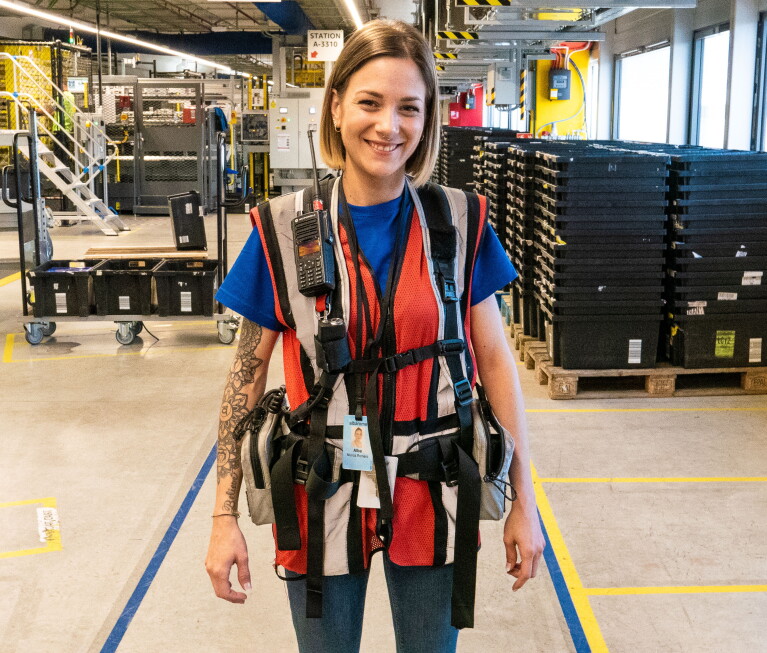Alba Muñoz, asociada en el área de robótica, con sus herramientas de trabajo. Sonriendo mirando a cámara, viste con su chaleco de trabajo lleno de bolsas llenas de utensilios. También viste con una camiseta azul y un pantalón tejano. De fondo está un pasillo del centro logístico del Prat de Llobregat. Castaña y con media melena tiene un piercing en la nariz. 