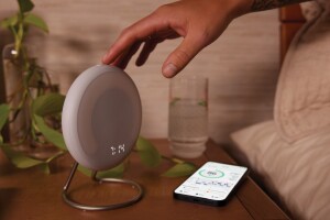 An image of Amazon's Halo Rise sitting on a wooden nightstand with a hand pressing a button from the bed. There is a phone, a glass of water, and a plant also sitting on the nightstand. 