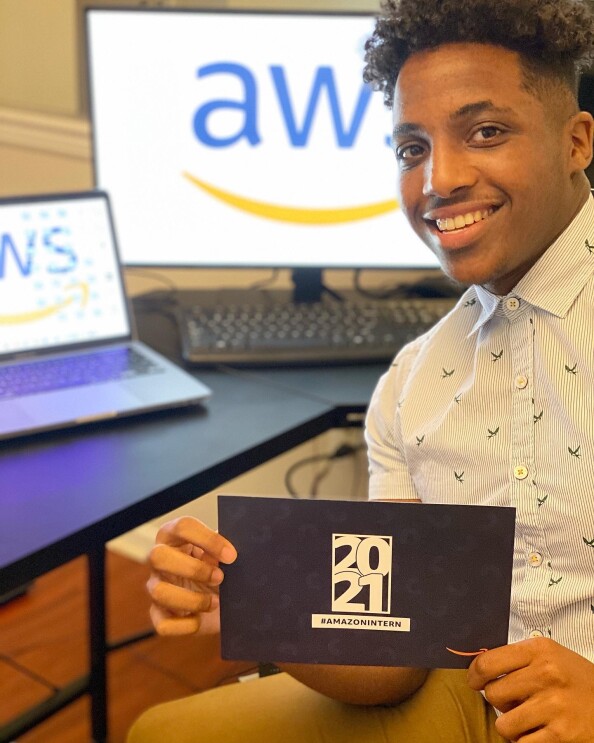 An image of a man smiling while sitting at a desk. He has two computer monitors in the background. both with the AWS logo on the screens. 