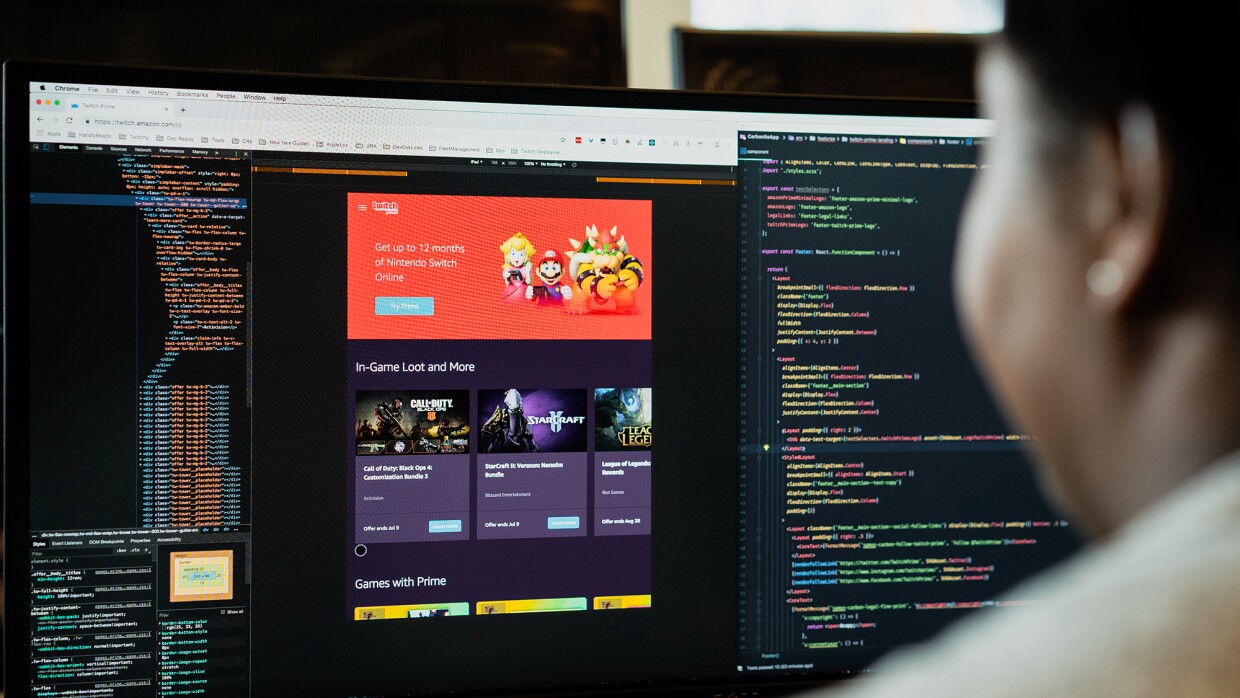 A woman sits at a computer, with two monitors in front of her. She is working within software, as illustrated by the lines of code displayed on the monitors.
