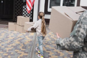 A child carries a moving box, behind her a man in military fatigues also carries a box. 