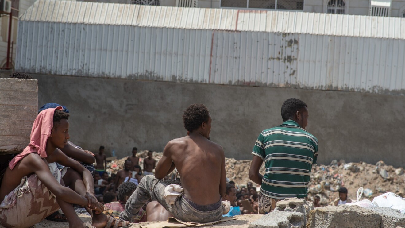 A group of male migrants sit together, looking at something outside of the camera's focus.