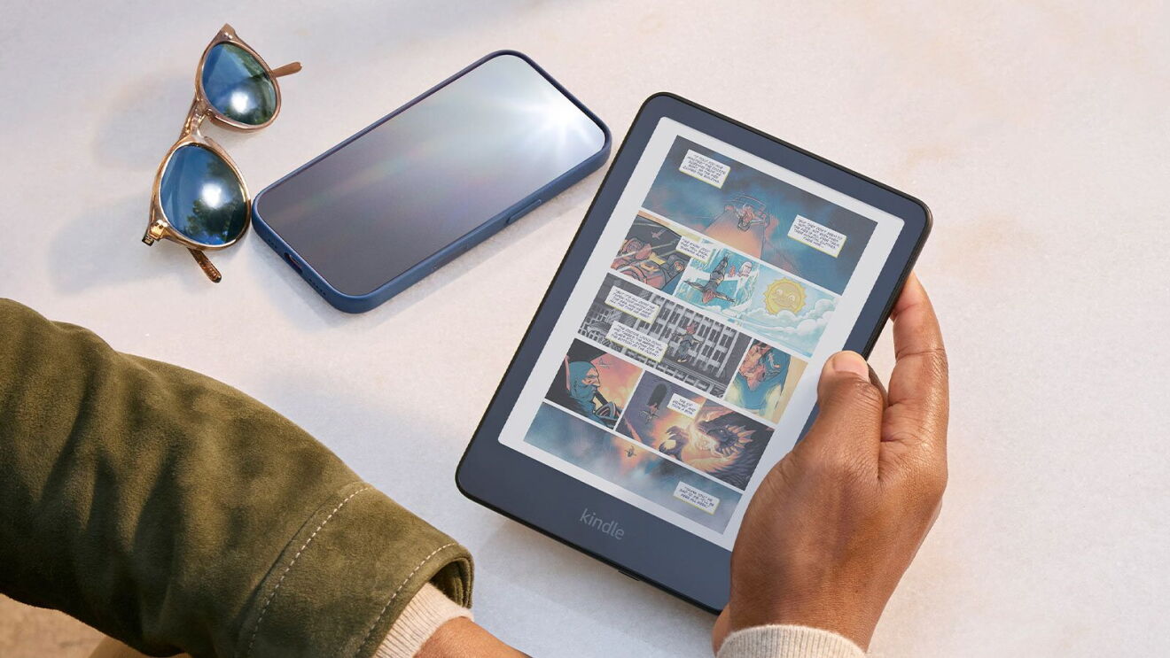 Person holding a Kindle e-reader displaying a colorful comic book page, with smartphone and sunglasses on white table