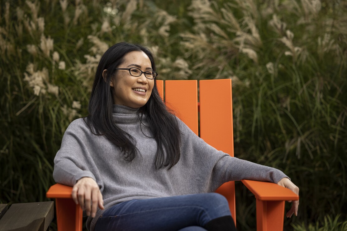 An image of a woman sitting in an orange chair outside. She is smiling and looking away from the camera and there is greenery behind her.